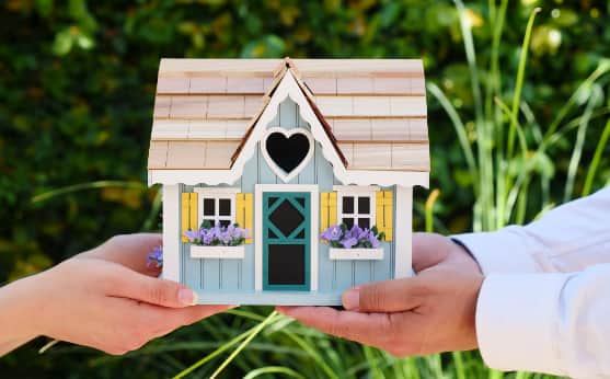 Zwei Hände tragen ein kleines Holzhaus (Dekoration) in der Hand. 