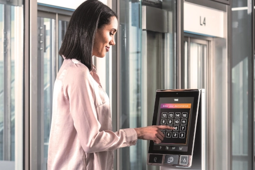 Eine Frau steht seitlich zum Aufzug im 4. Stockwerk und drückt auf einem Touchpad die Taste 6.