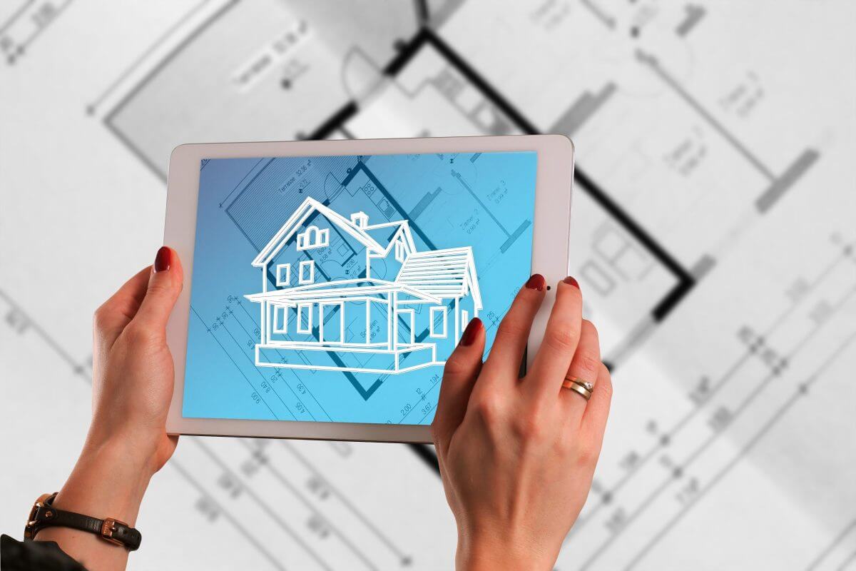Frau hält ein Tablet in den Händen, auf dem ein 3D-Haus zu sehen ist und im Hintergrund ein analoger Bauplan.