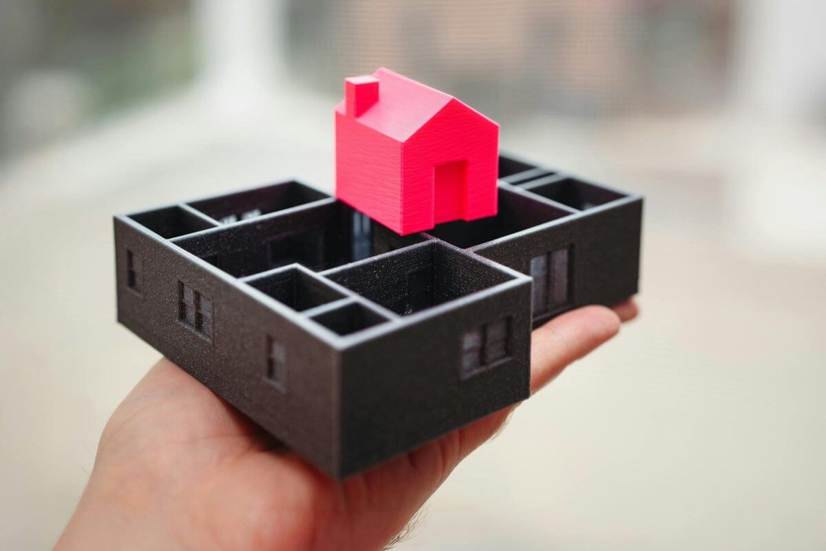 Eine Hand hält ein 3D-gedrucktes Modell eines Hauses mit roten Wänden und schwarzem Grundriss.