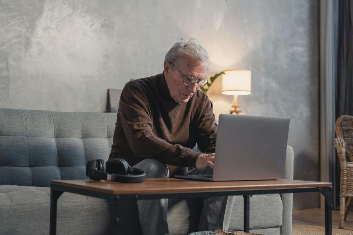 Älterer Mann mit Brille sitzt konzentriert am Laptop auf einem grauen Sofa im Wohnzimmer.